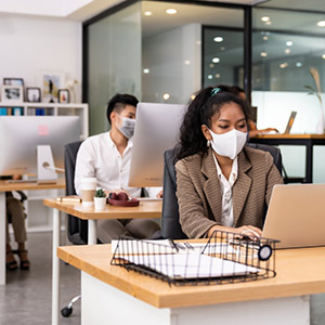 Des collègues de travail portant un masque assis à leur bureau et utilisant des ordinateurs.